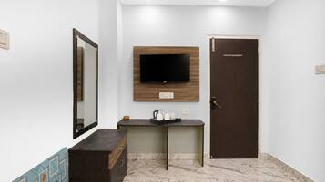 Ein Hotelzimmer mit Flachbildfernseher an der Wand, einer Holztür, einem kleinen Schreibtisch mit Wasserkocher und einem Spiegel.