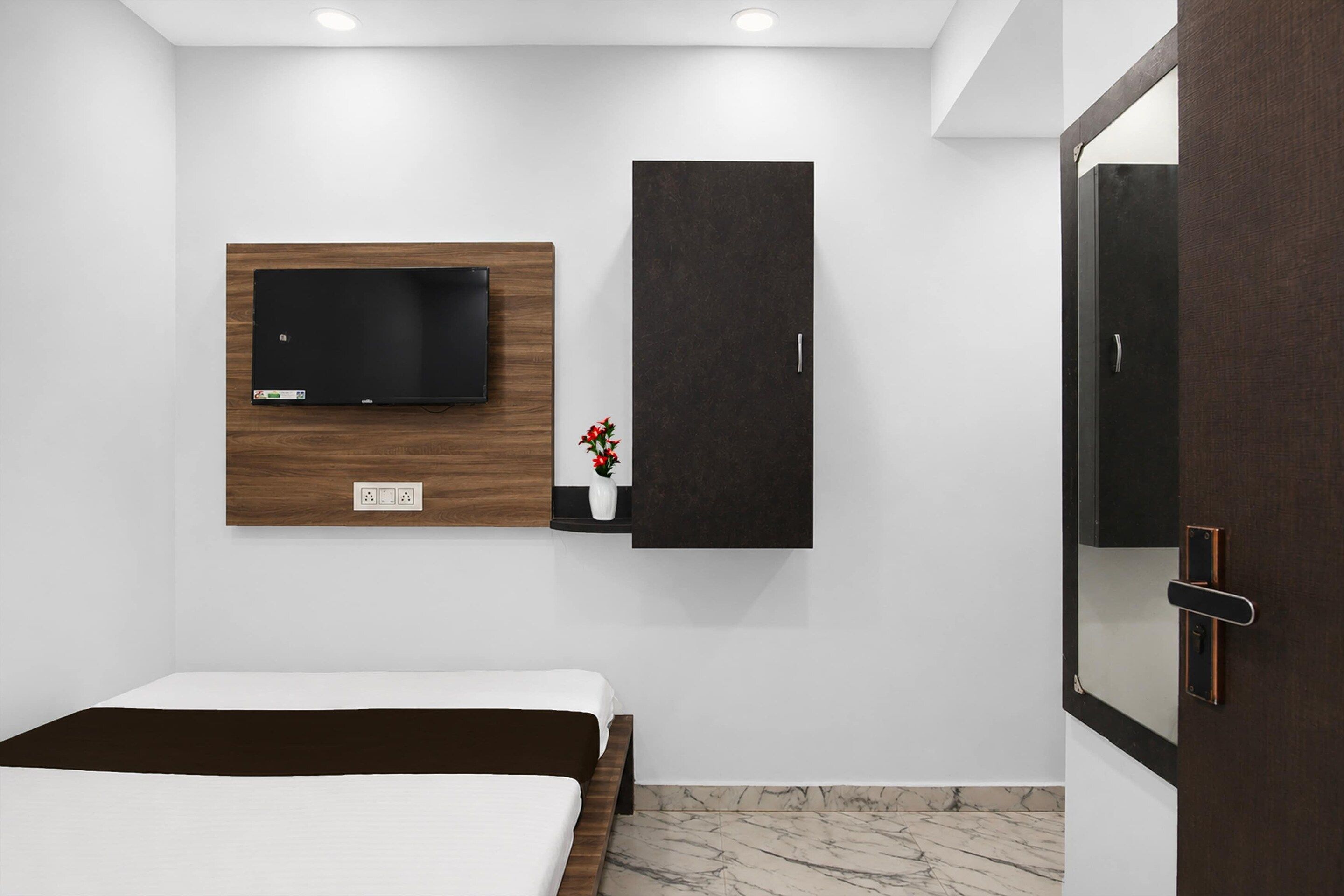 A bedroom with a bed, a wall-mounted TV, a dark cabinet, and a door with a mirror.