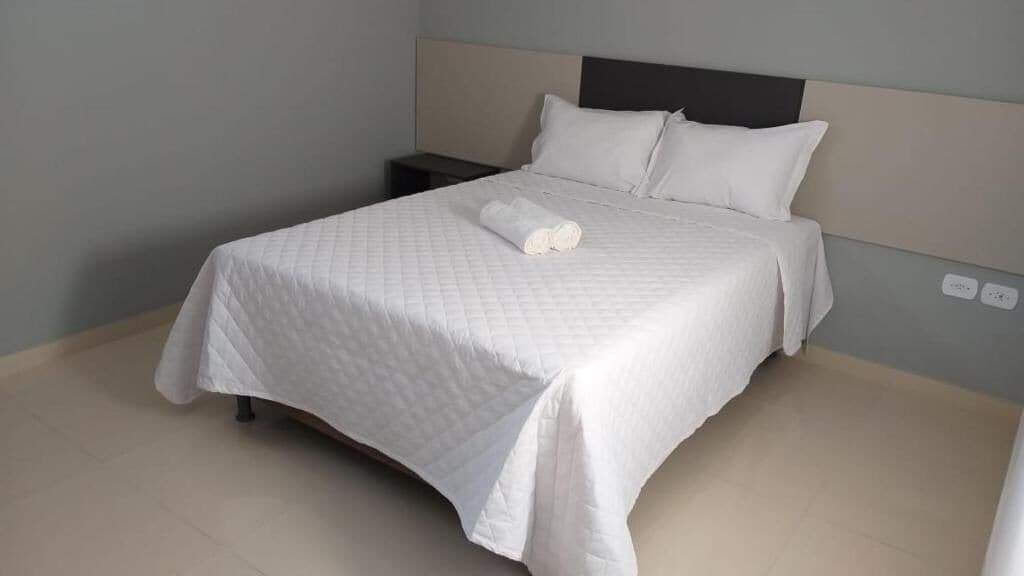 A neatly made bed with a white quilted bedspread and pillows, a bedside table with a lamp, and a wall socket.
