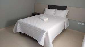 Una cama bien hecha con una colcha blanca acolchada y almohadas, una mesita de noche con una lámpara y un enchufe de pared.