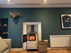 A room with a wood-burning stove, a framed Peter Vardy t-shirt on the wall, and a woven basket on the floor. - Eldon Cottage, Corrie, Isle of Arran (Corrie, Isle of Arran)