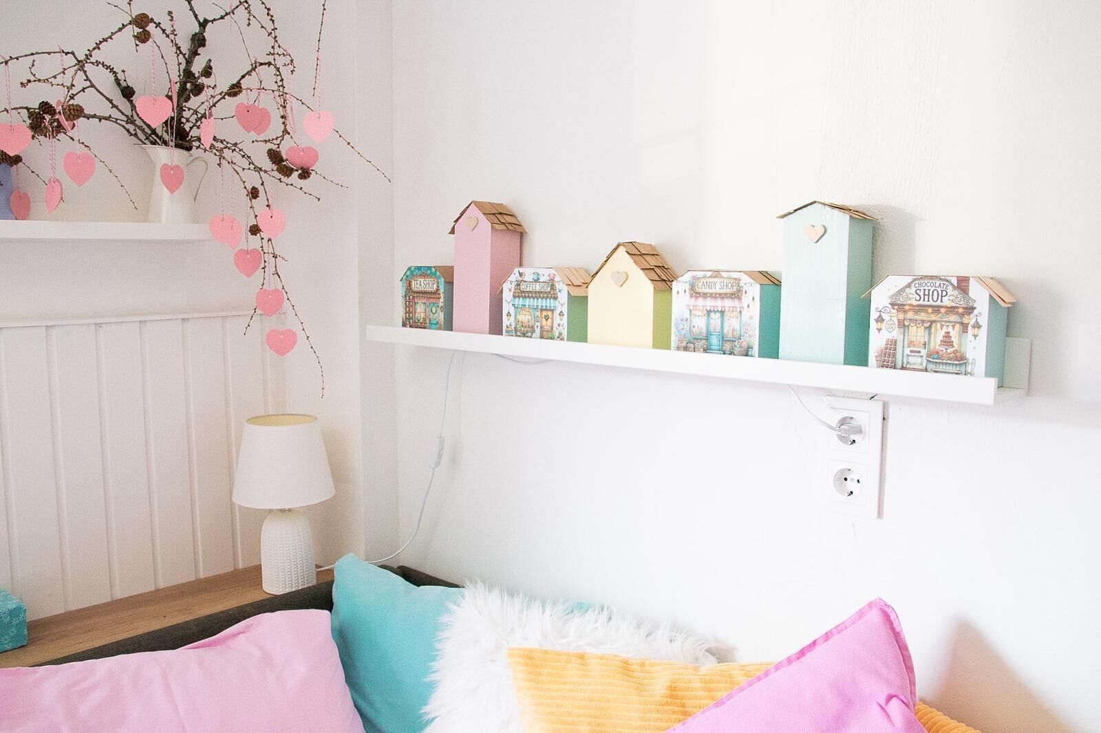 A bedroom with a bed, pillows, a bedside lamp, a wall-mounted shelf with decorative houses, and a wall-mounted power outlet.