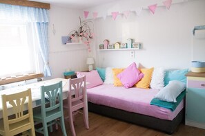 A cozy room with a bed, a table, chairs, and a shelf with decorative items.
