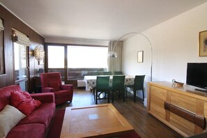 Ett vardagsrum med en röd soffa, ett träbord som soffbord, en matplats med ett bord och stolar, en tv-apparat på ett träskåp och ett stort fönster med gardiner.