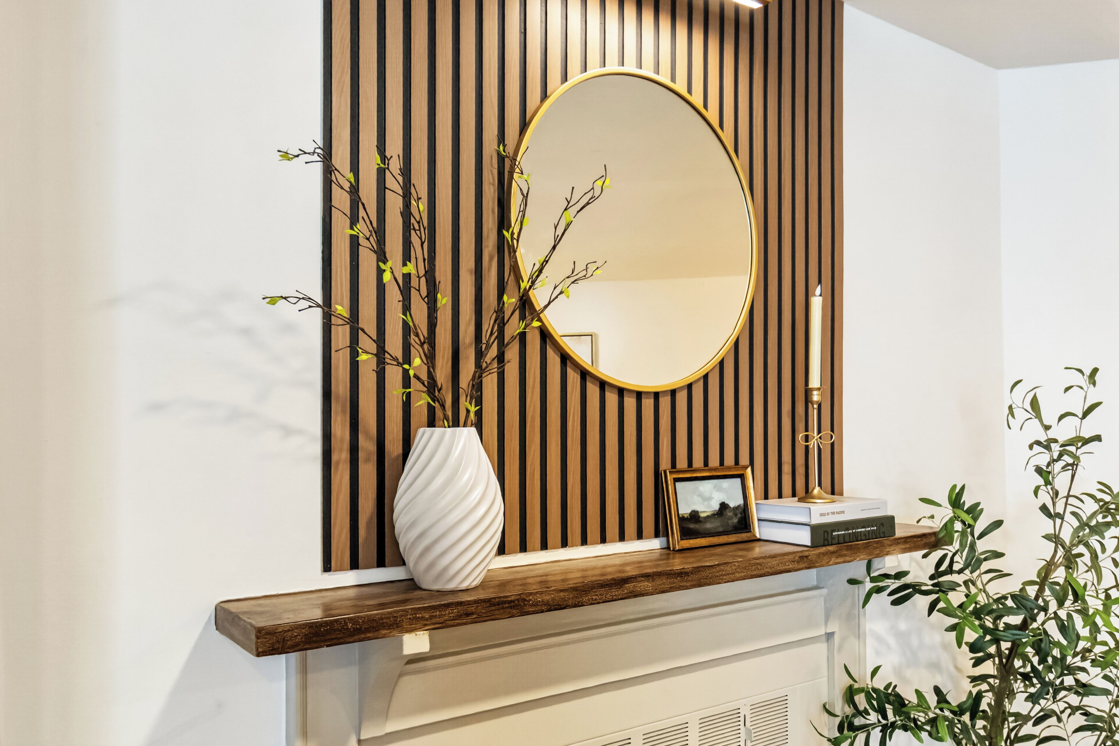 Un intérieur moderne avec un mur lambrissé en bois, un miroir rond, un vase blanc orné de branches, un tableau encadré et un livre posé sur une cheminée.