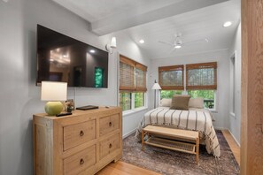 Quarto com cama, cómoda de madeira, televisão na parede e janelas com persianas.