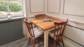 Een zithoek met een eettafel en stoelen, een radiator en een raam met uitzicht op gebouwen en groen.