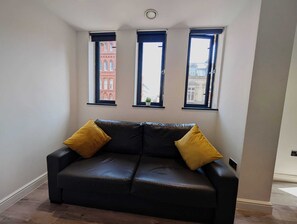 Ein Zimmer mit einer schwarzen Ledersofa, zwei Fenstern mit schwarzen Rahmen und Blick auf Gebäude.