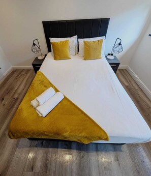 Ein Bett mit schwarzem Kopfteil, weißer Bettwäsche und zwei gelben Kissen. Zwei Nachttische mit geometrischen Lampen stehen daneben. Eine senffarbene Tagesdecke und zwei aufgerollte weiße Handtücher liegen auf dem Bett.