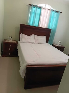 A bedroom with a wooden bed, two nightstands, a window with curtains, and a small table with a flower. - Delightful 2-bedroom house with AC in Puerto Plata (Puerto Plata)