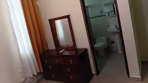 Ein Zimmer mit einem hölzernen Schminktisch, einem Spiegel und einem durchgehenden Türbogen sichtbaren Badezimmer.