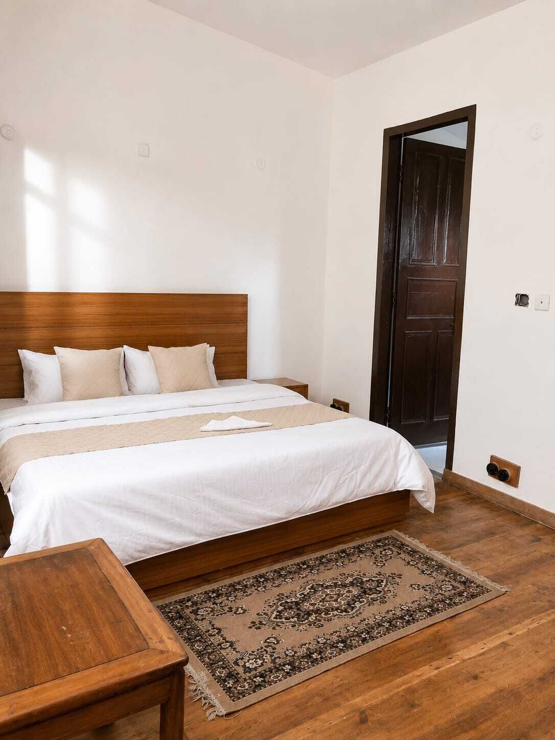 Quarto com cabeceira de madeira, uma cama grande com roupa de cama branca, uma pequena mesa de madeira e um tapete estampado.