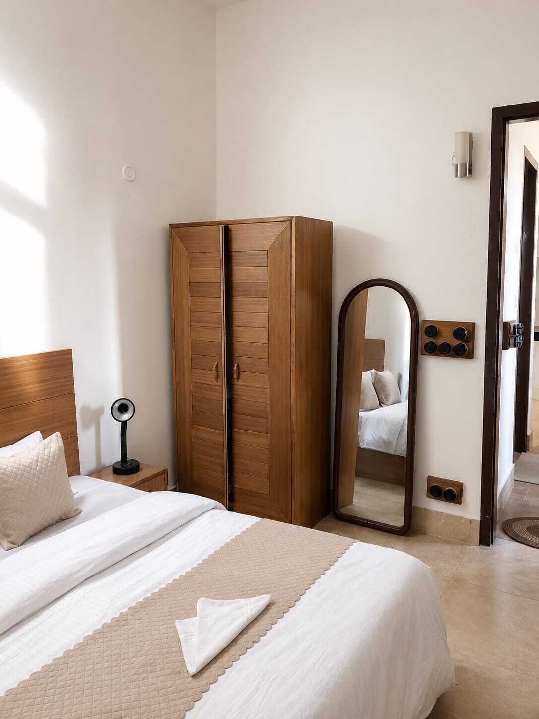 A hotel room with a bed, a wardrobe, a bedside table with a lamp, a mirror, and a door with a handle.