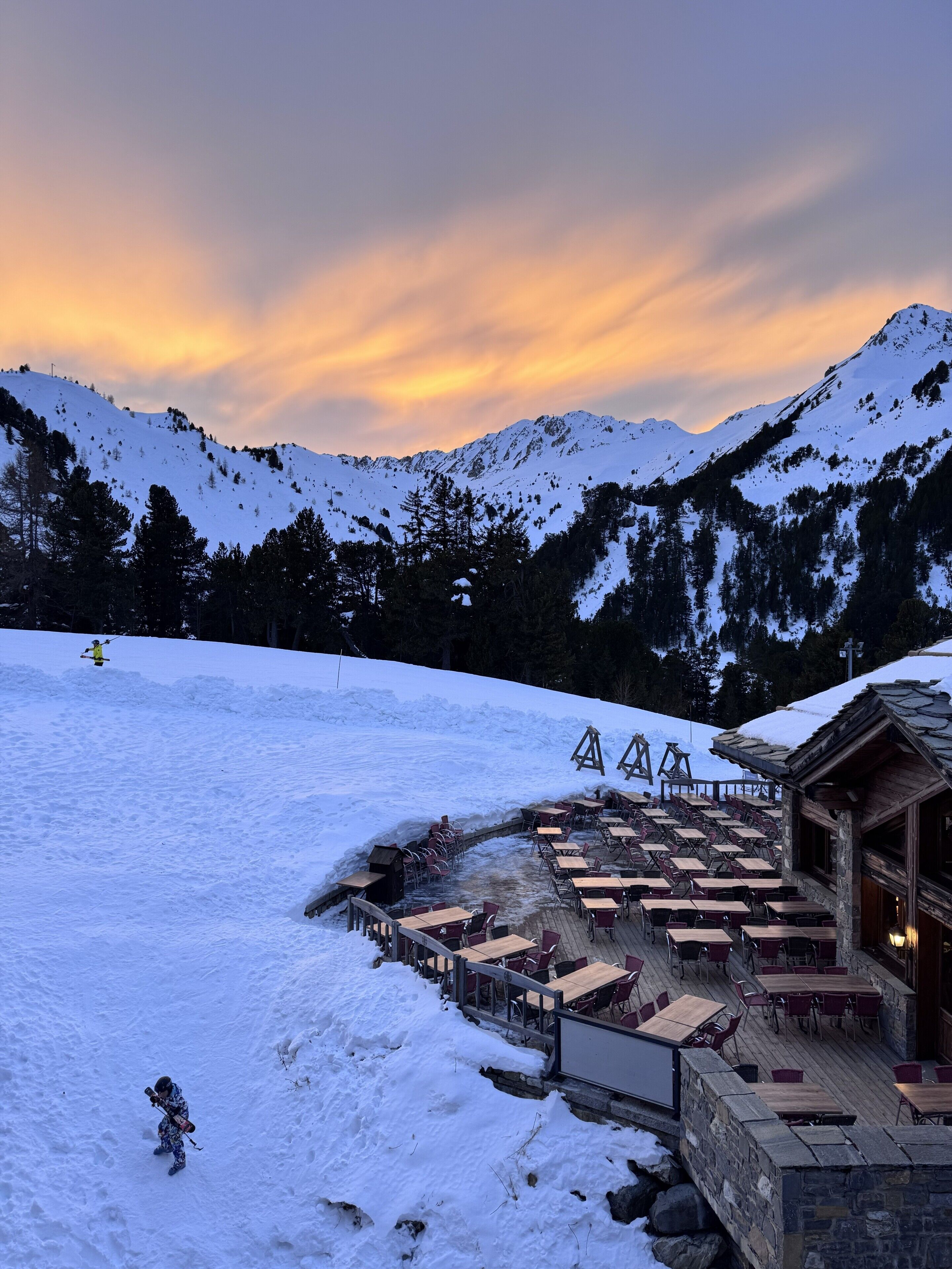 A snow-covered outdoor seating area with tables and chairs, a wooden building with a sloped roof, and a mountainous backdrop.