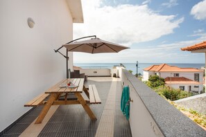Eine Terrasse mit Holztisch und Sonnenschirm, mit Blick auf die Küste.