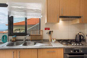 A kitchen with a double sink, wooden cabinets, a gas stove, and a view of the sea through a window. - Casa D'Avó Apartment with Sea View and Private Terrace (Santa Cruz)