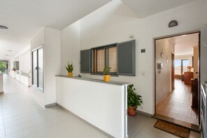 Ein Hotelgang mit einer Rezeption, Topfpflanzen, einem Fenster mit Rollläden und einer Tür zu einem Zimmer.