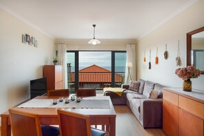 A modern living room with a dining area, large windows, a view of the sea, and a balcony with a red-tiled roof. - Casa D'Avó Apartment with Sea View and Private Terrace (Santa Cruz)
