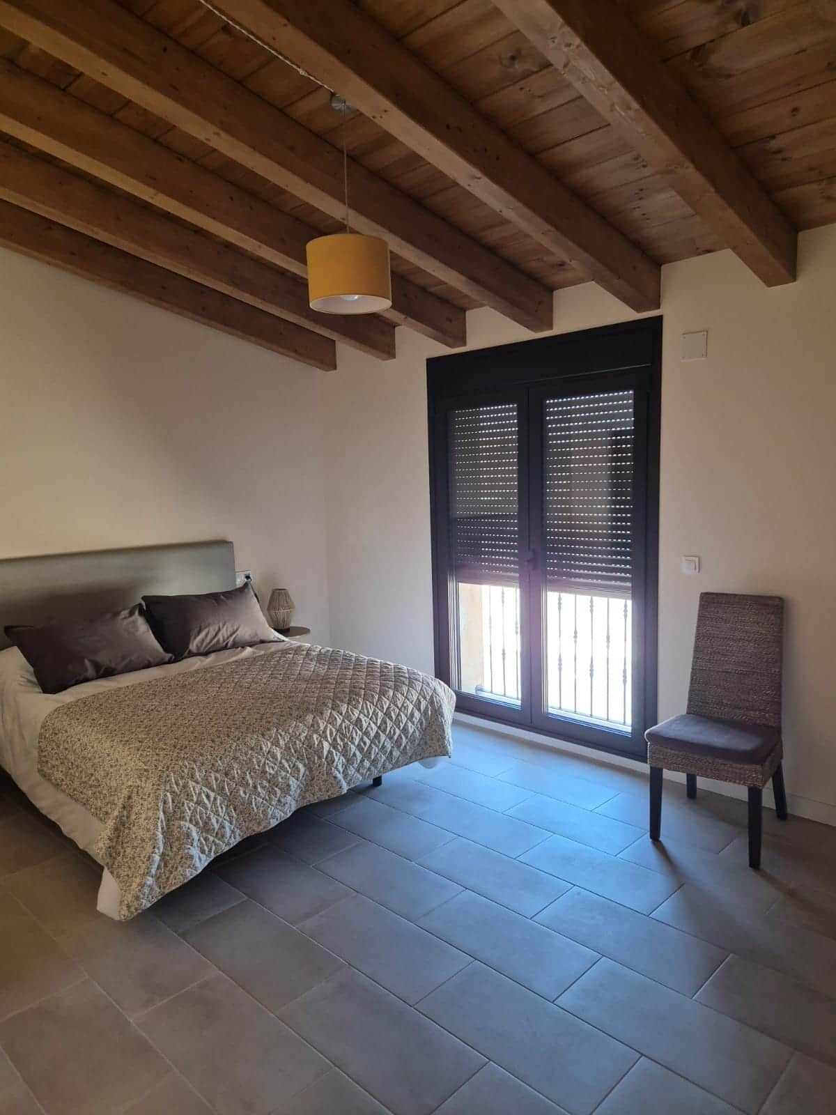 A bedroom with a bed, a chair, a window with blinds, and a wooden ceiling.