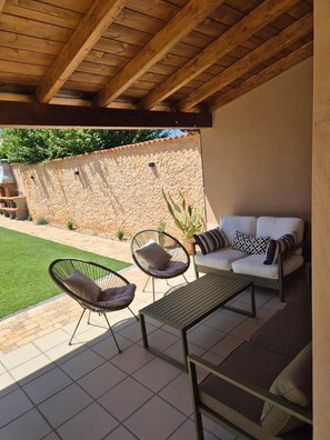 An outdoor patio area with a wooden ceiling, a stone wall, a white sofa, two chairs, and a table.