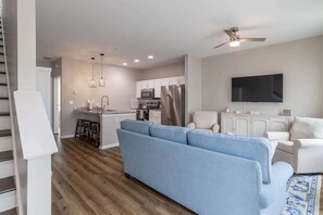 A modern living room with a blue sofa, a television, and a kitchen area with white cabinets and appliances. - The Dolly (Oxford)