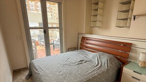 Ein Schlafzimmer mit Bett, Nachttisch, Lampe, Deckenleuchte und einem Fenster mit Blick auf Gebäude.