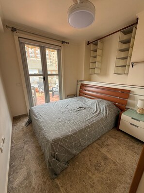 Ein Schlafzimmer mit Bett, Nachttisch, Lampe, Deckenleuchte und einem Fenster mit Blick auf Gebäude.