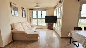 Ein geräumiges Wohnzimmer mit einem beigefarbenen Sofa, einem Flachbildfernseher an einem Holzbodenständchen und einem Esstisch mit gemustertem Tischlauf.