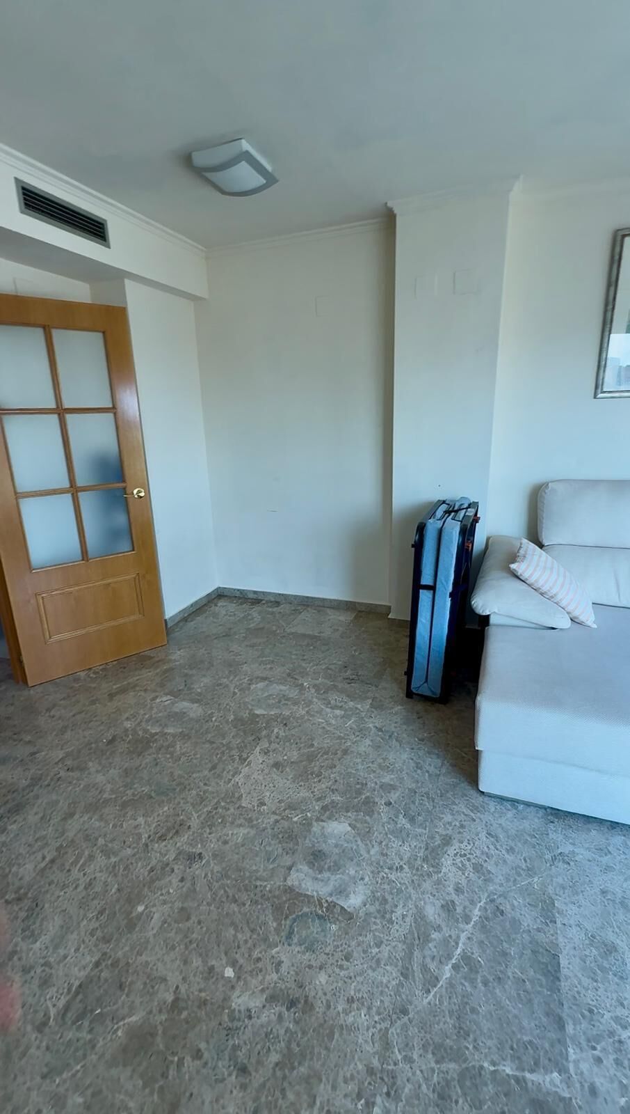 Ein Zimmer mit einem weißen Sofa, einer Holztür mit Glasscheiben, einem Lüftungsgitter an der Decke und einem Stapel gefalteter Kleidung.
