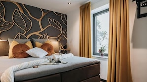 Et moderne soveværelse med en stor seng, et vægdekor med bladmotiver, et vindue med gardiner og et natbord med en lampe.