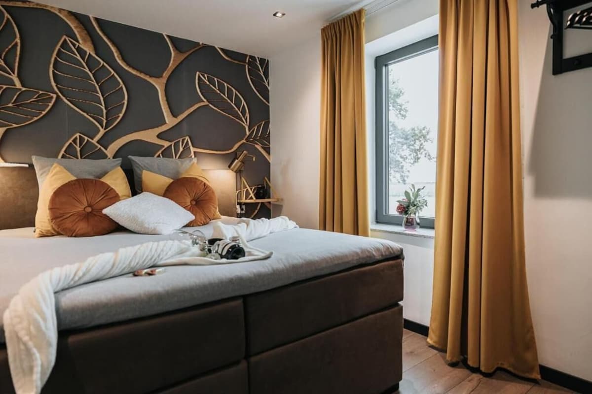 Ein modernes Schlafzimmer mit einem großen Bett, einer Wand mit dekorativem Blattmuster, einem Fenster mit Vorhängen und einem Nachttisch mit Lampe.