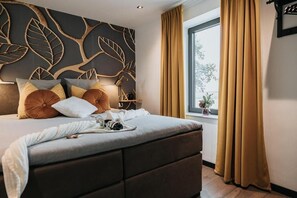 Ein modernes Schlafzimmer mit einem großen Bett, einer Wand mit dekorativem Blattmuster, einem Fenster mit Vorhängen und einem Nachttisch mit Lampe.
