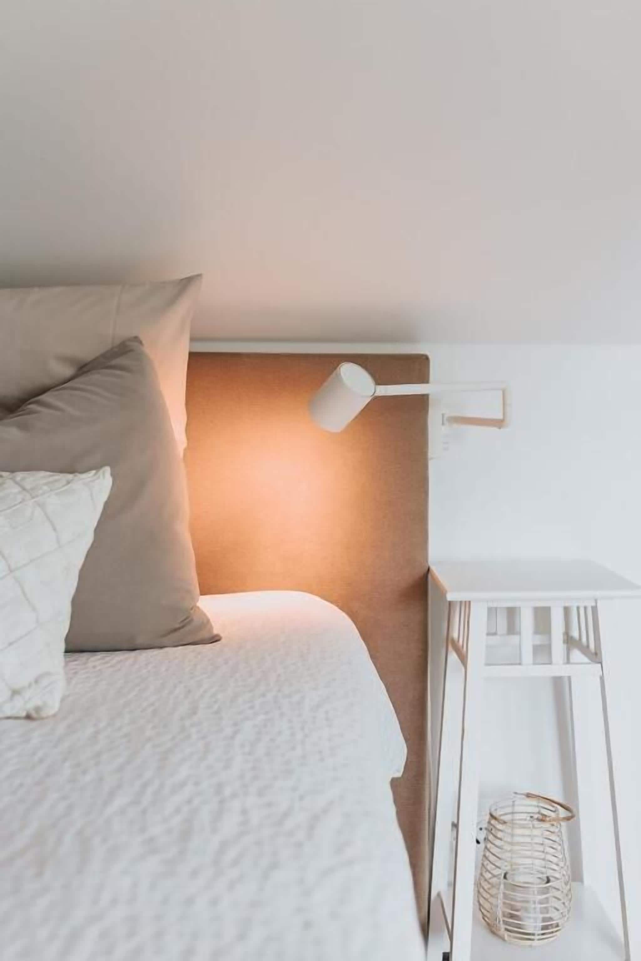 A bedroom with a bed, pillows, a bedside lamp, a white bedside table, and a wicker basket.