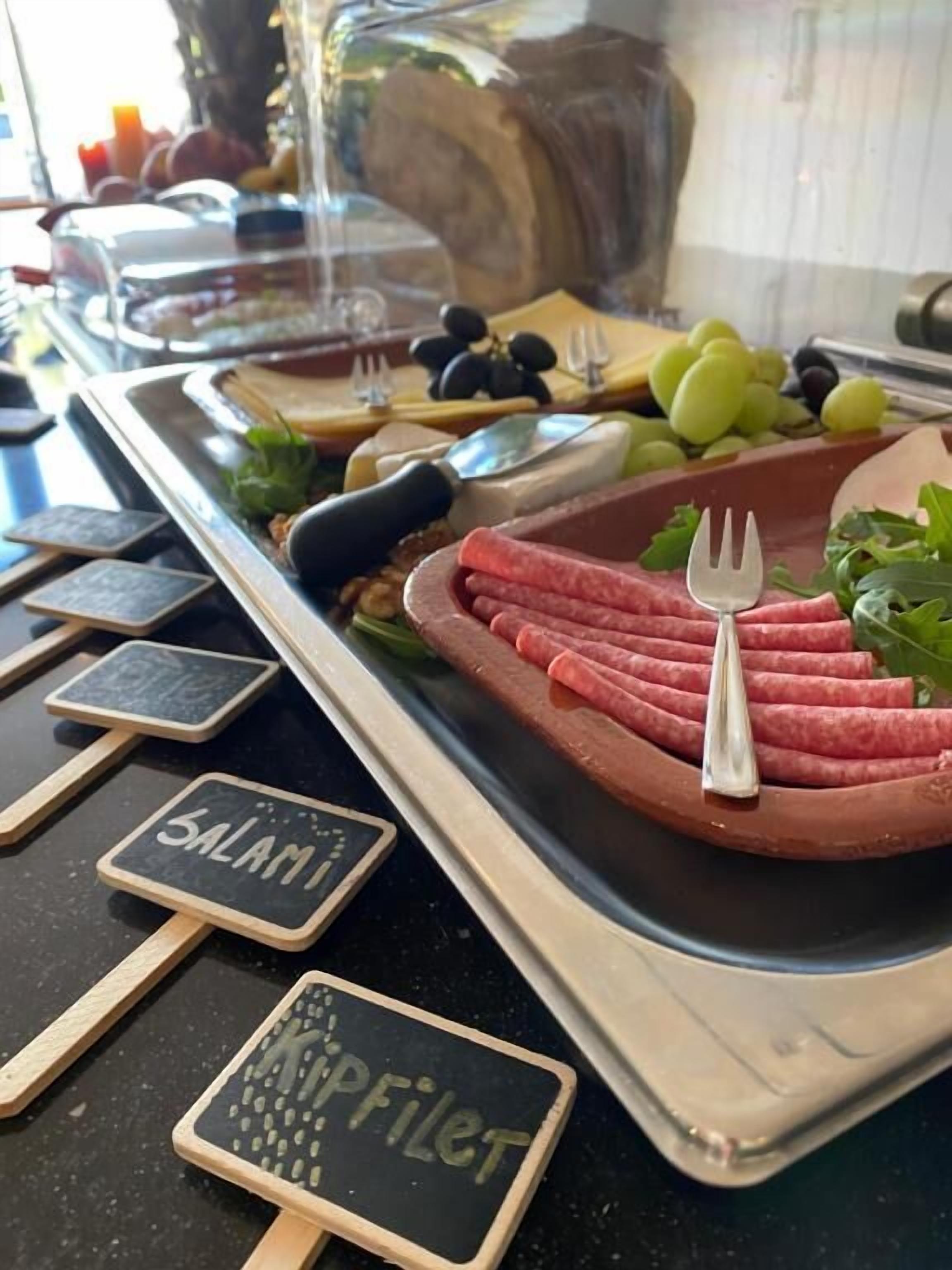 Un bufé con diversos alimentos, incluyendo embutidos en rodajas y uvas, con etiquetas que dicen 'SALAMI' y 'KIPFILET'.