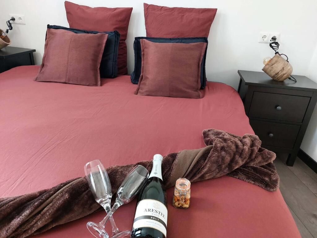 Ein Bett mit roter Bettdecke, zwei Kissen, eine Weinflasche, zwei Gläser und eine Nussmischung auf einer braunen Decke.