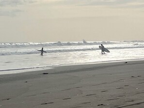 A beach with two individuals carrying surfboards, a sandy shore, and waves in the background. - Spacious 5-bedroom Front beach house in Pochomil (Pochomil)