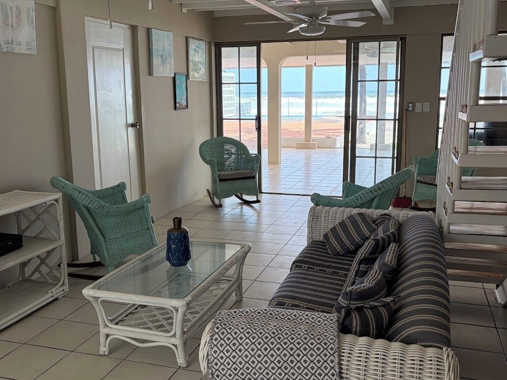 A living room with a glass coffee table, a sofa, and a chair. There is a view of the beach through large glass doors.