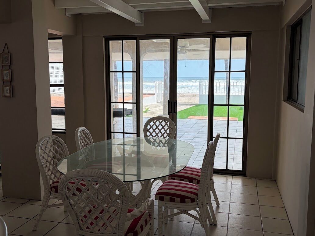 A dining area with a glass table and wicker chairs, large sliding glass doors leading to a patio with a view of the ocean.