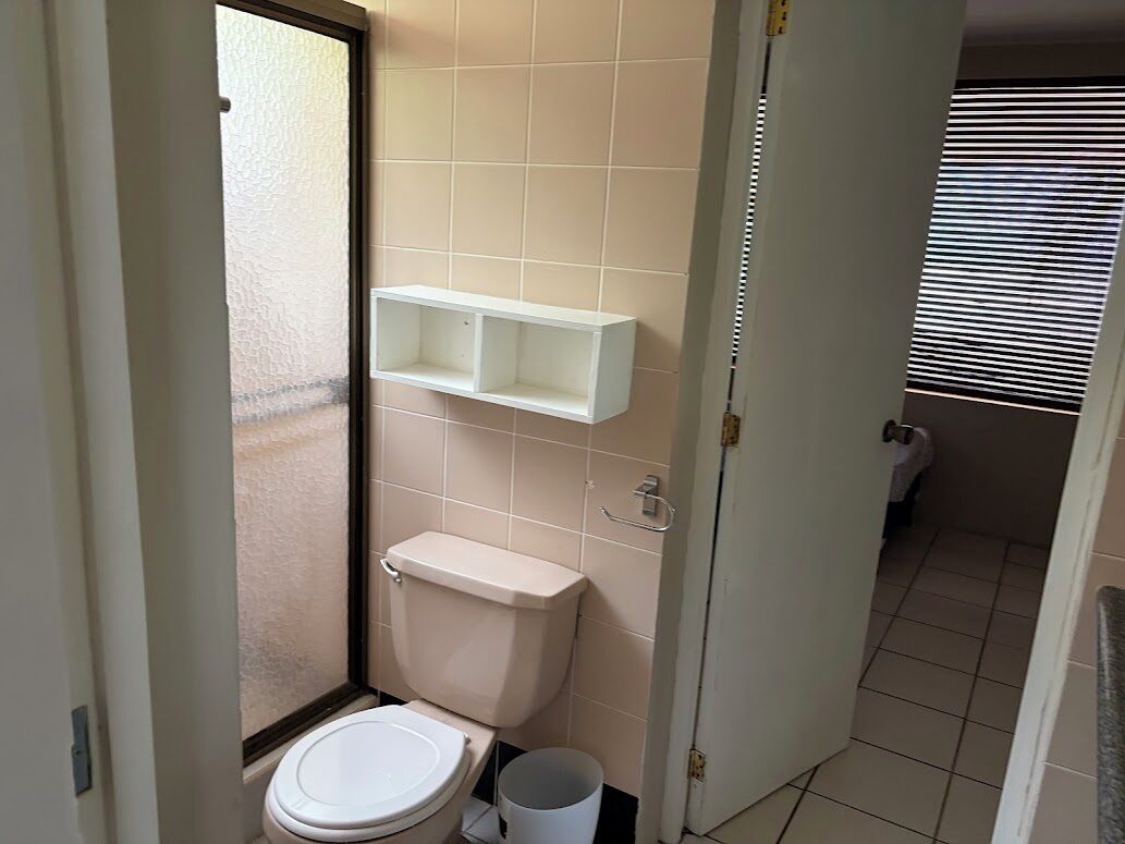 Un baño pequeño con inodoro, un estante blanco y una ventana con persianas.