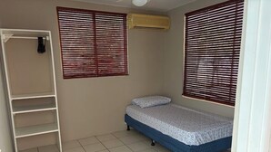 A small, single-bed room with a wardrobe, two windows with blinds, and an air conditioning unit.