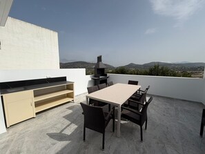Eine Dachterrasse mit Esstisch, Stühlen, einem eingebauten Grill und Blick auf Stadt und Berge.