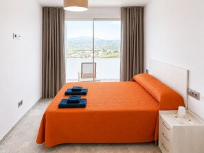 Ein Hotelzimmer mit einem großen Bett, einem Nachttisch mit Lampe und Blick auf die Stadt durch ein großes Fenster.