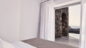 A bedroom with a bed, white curtains, and a view of a stone wall and ocean. - Villa Lythion, Divine Sea Views, Private Pool, Furnished Terrace, Sanrotini (Oia, Santorini)