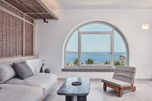 A modern living room with a large arched window, a sofa, a chair, and a small table. - Corvionis Suite, Divine Sea Views, Private Pool, Furnished Terrace, Sanrotini (Oia, Santorini)