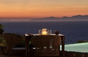 A table set for two with a candle, two glasses, and a decorative item, overlooking a body of water and distant mountains. - Corvionis Suite, Divine Sea Views, Private Pool, Furnished Terrace, Sanrotini (Oia, Santorini)