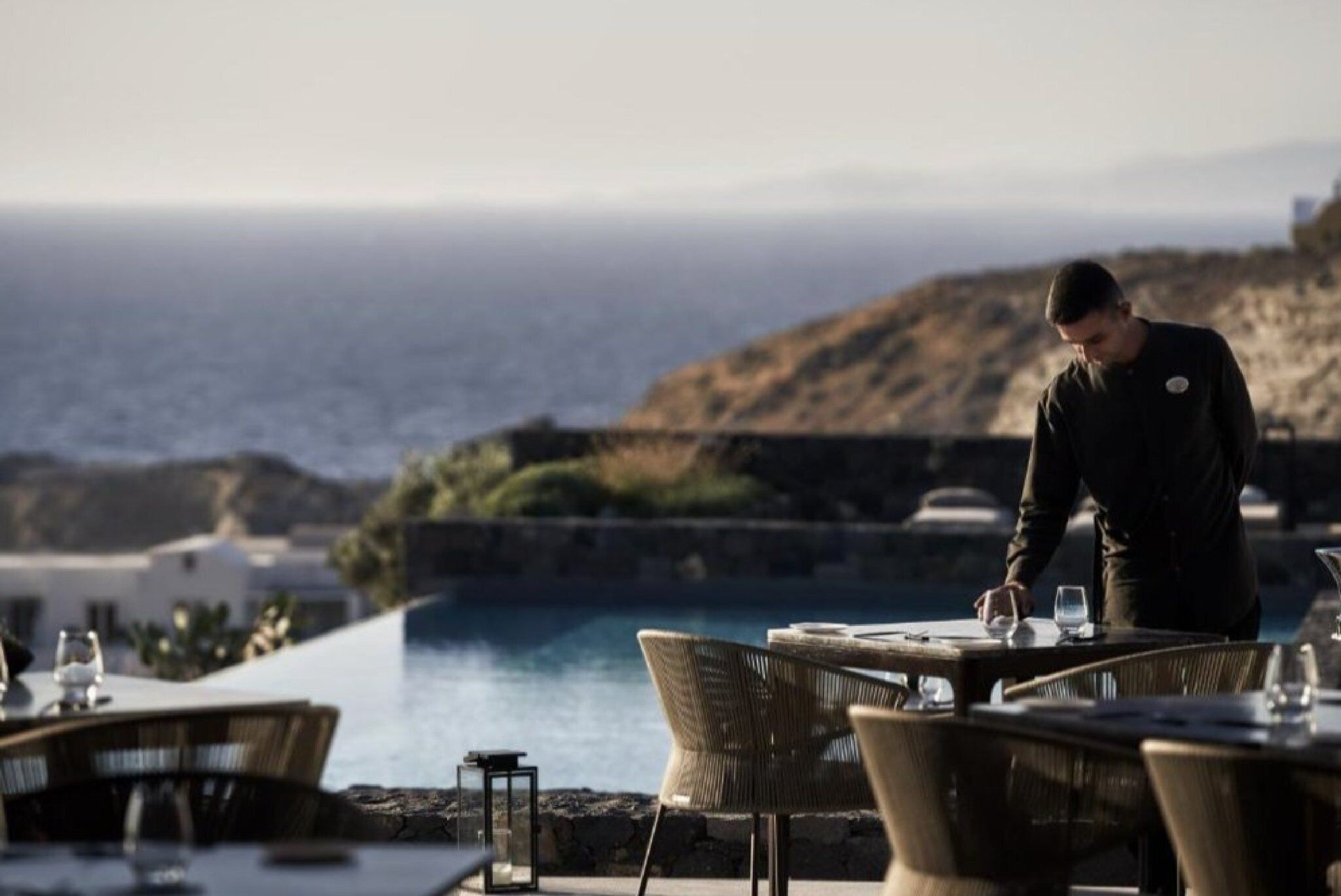 A dining area with tables and chairs overlooking a body of water, with a person attending to the table.