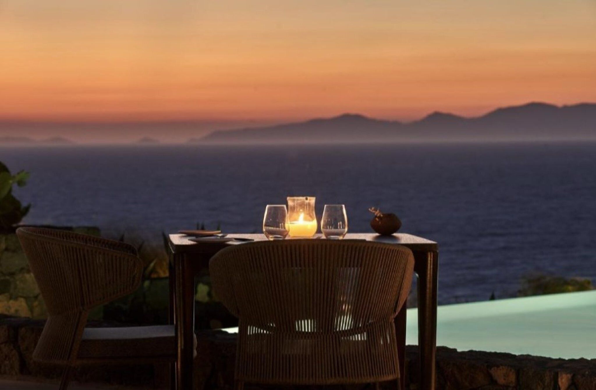 Mesa para dos con vela, dos vasos y un objeto decorativo pequeño, con vista a un cuerpo de agua y montañas al fondo.