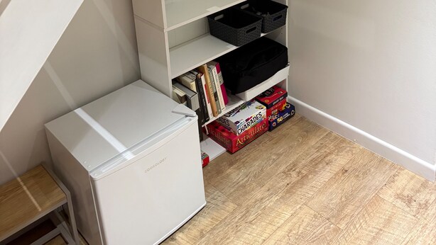 A compact room with a white mini-fridge, a wooden floor, and a white shelving unit containing various items. - Tranquil Matlock Bath Retreat With 2 Bedrooms (Matlock)