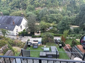 Ein Wohnhaus mit umzäunter Garten, einer Terrasse mit Sitzgelegenheit und Blick auf einen bewaldeten Hügel.