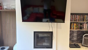 A flat-screen TV mounted on a wall, a framed picture below it, a small shelf with decorative items, and a bookshelf with DVDs. - Tranquil Matlock Bath Retreat With 2 Bedrooms (Matlock)